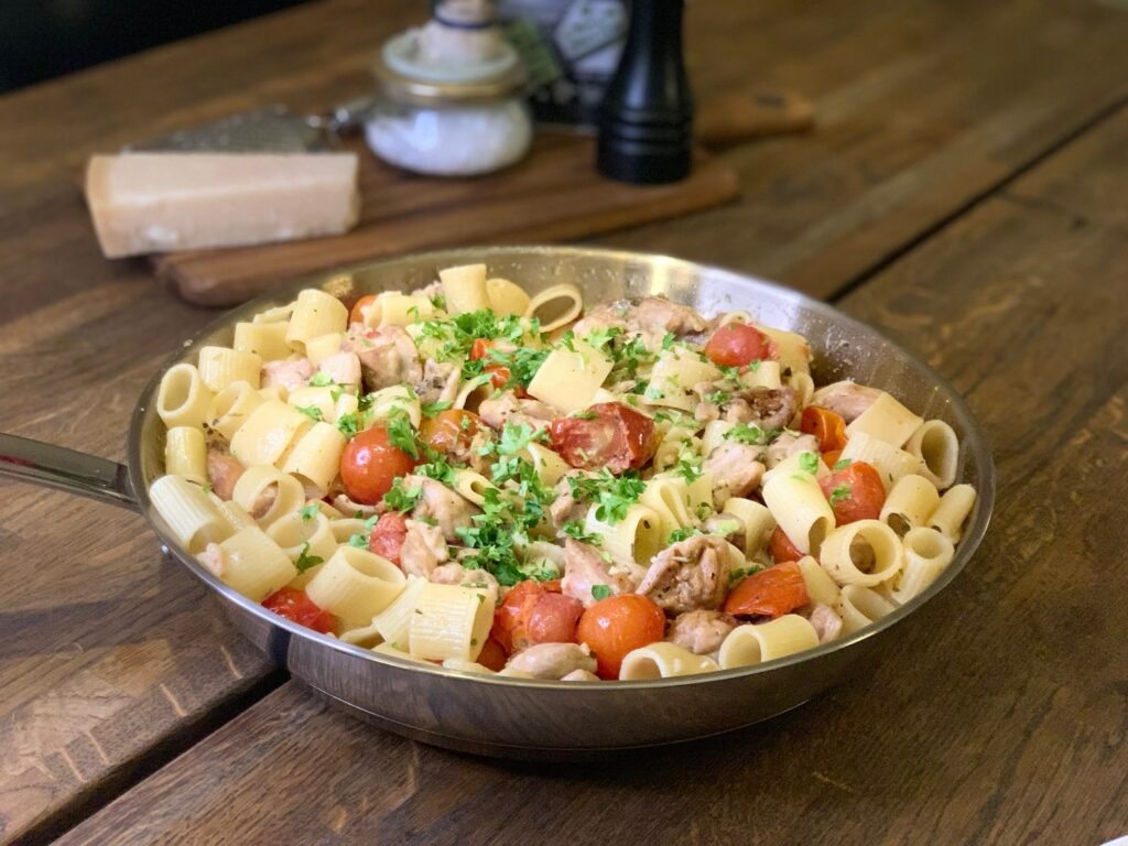 Pasta med kyckling och tomater i en stekpanna på ett brunt bord.