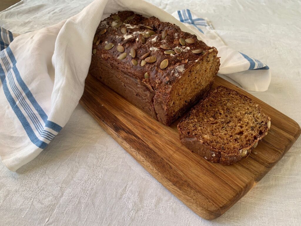 En brun limpa och en skiva av limpan som ligger på en brun skärbräda med en vit bakduk på bakre delen av limpan.