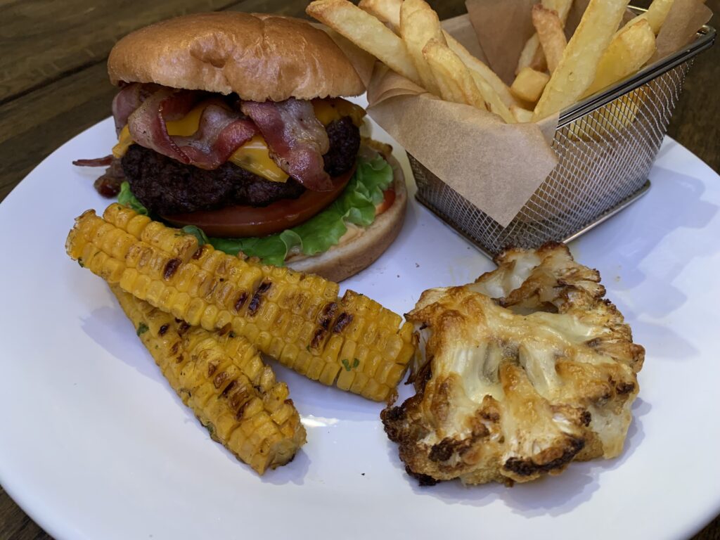 Hamburgare med bröd, pommes, grillad majs och gratinerad blomkål på vit tallrik