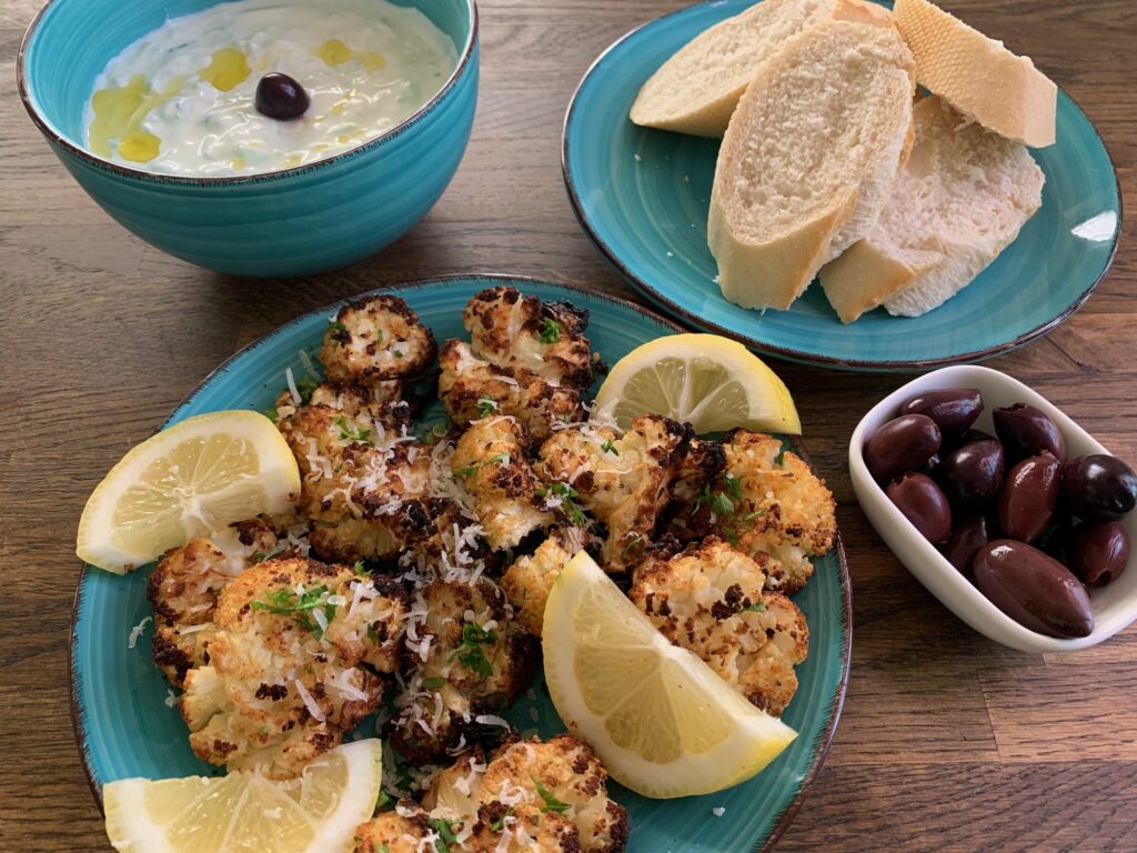 Grön tallrik med ungsgratinerad blomkål med citronskivor, en grön tallrik med bröd, en skål med vit dipp och en vit liten skål med svarta oliver.