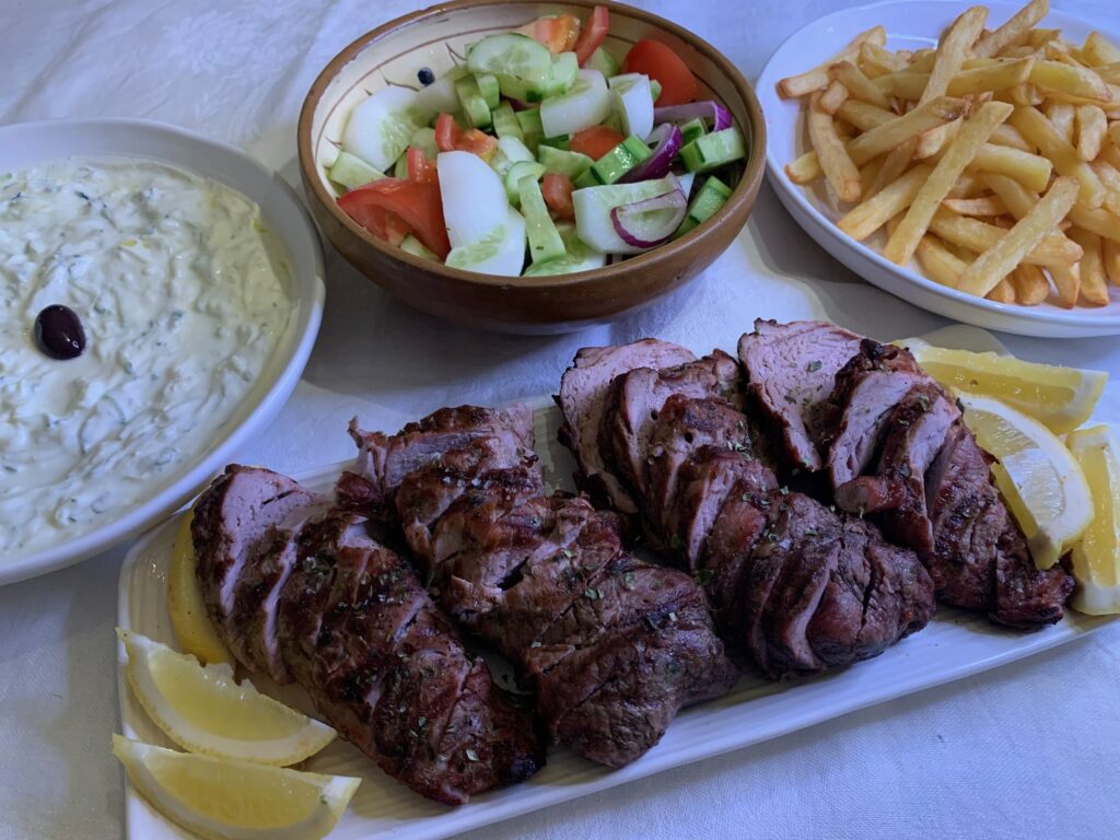 Helgrillad fläskfilé som ligger uppskivat på ett av långt vitt fat med citronklyftor. I bakgrunden en vit djup tallrik med tzatziki, en terracottafärgad skål med sallad och en vit djul