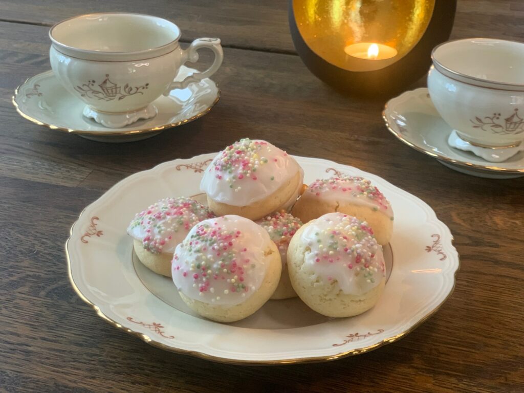 Italienska julkakor med glasyr toppade med strössel på ett vitt fat. I bakgrunden två kaffekoppar på tallrikar och ett värmeljus i svart och guldfärgad lykta.