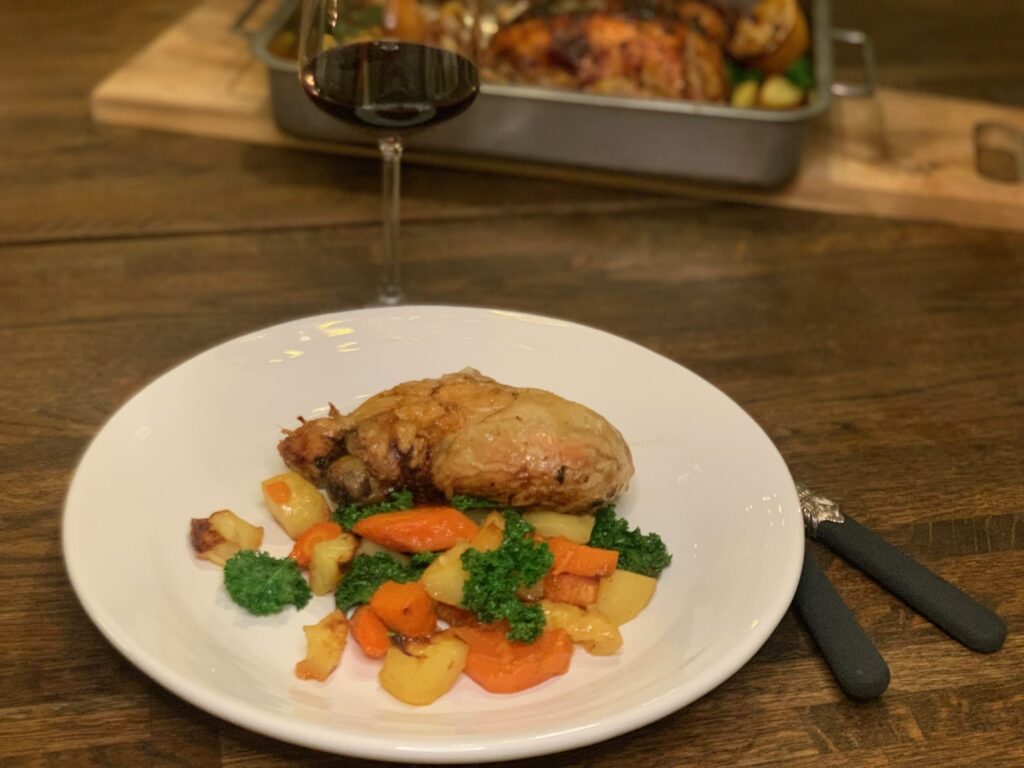 Rosta rotfrukter i härliga färger och kyckling på vit tallrik. I bakgrunden ett glas rött vin och den ugnsfasta formen med kyckling och rotfrukter.