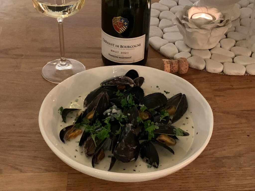 Moules marinières, musslor kokata i vitt vin och grädde i en djup vit tallrik. i bakgrunden en flaska, ett vinglas och ett ljus.