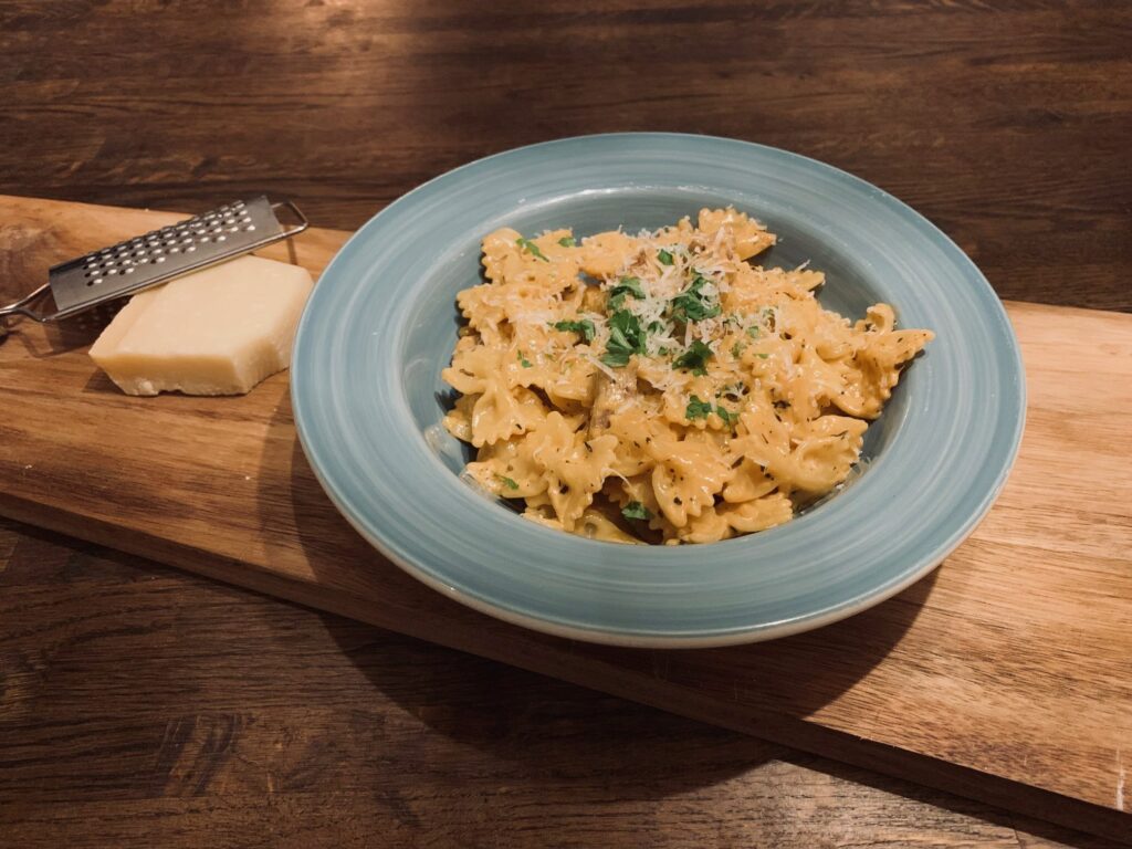 Pasta som ser ut som rosetter i en ljusgrön skål som står på en träbräda. Till vänster ligger en bit hårdost och ett rivjärn