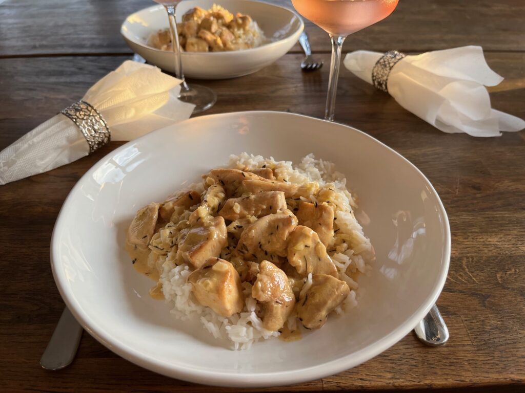 Kycklinggryta och ris på vit djup tallrik, tillvänster en servett i servettring och ovanför ett vinglas med rosa vätska.